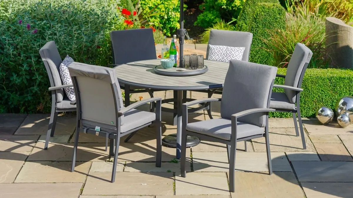 Garden patio with round dining table, six grey padded chairs, wine bottle and glassware, surrounded by flowering plants and hedges.