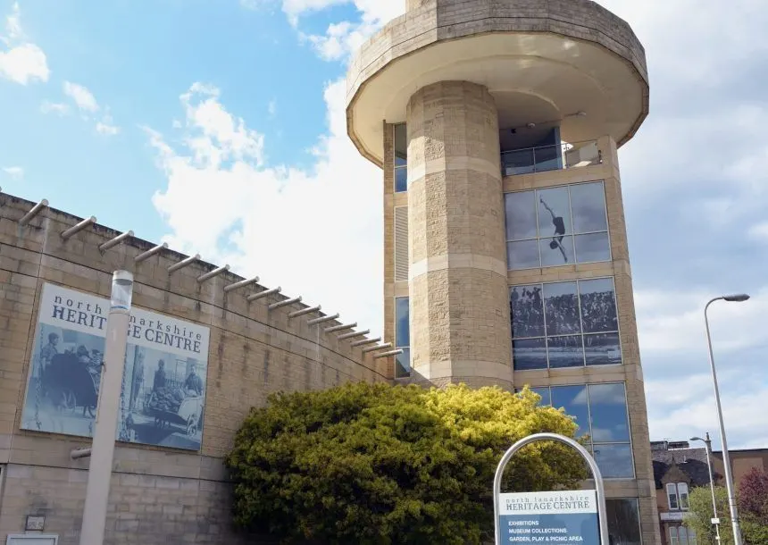A cylindrical stone tower with large windows beside a single-storey stone building with blue banners, set against a blue sky with clouds.