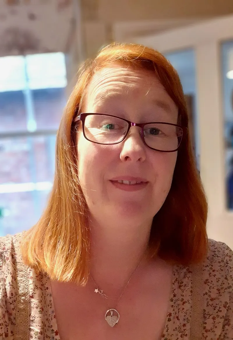 Woman with red hair and glasses smiling at the camera indoors near windows.