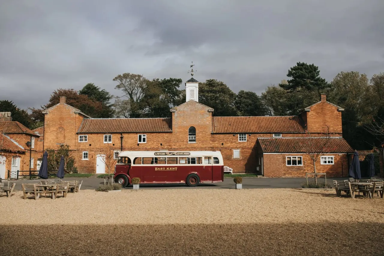 Vintage red bus parked in front of a brick building with a bell tower, on a sandy beach with wooden seating.