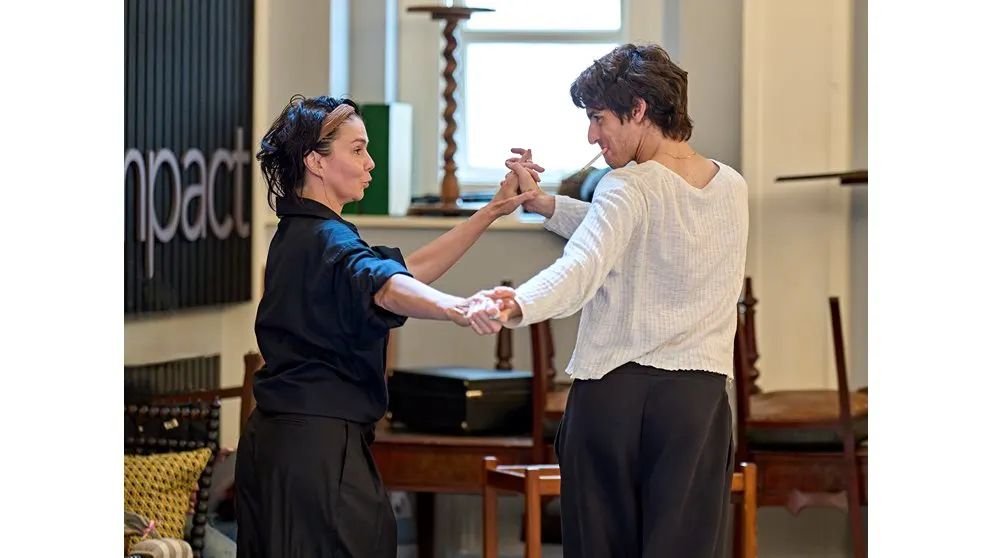 Two women holding hands while dancing together in a theatre interior with wooden furniture and period décor.