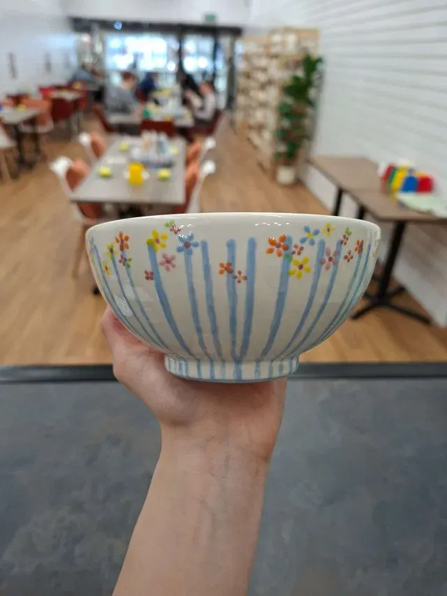 Hand holding a decorative ceramic bowl with blue stripes and coloured flower patterns in an art studio space.