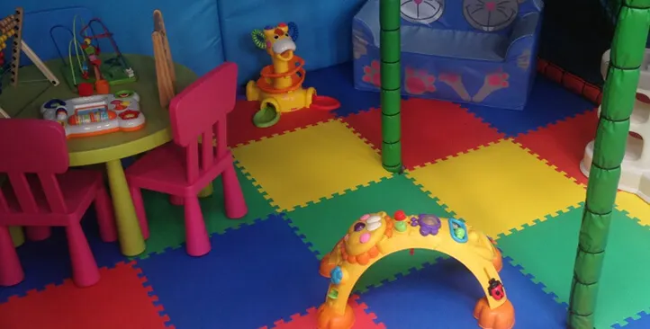 Colourful soft play area with foam floor mats, toy furniture, and play structures for young children.