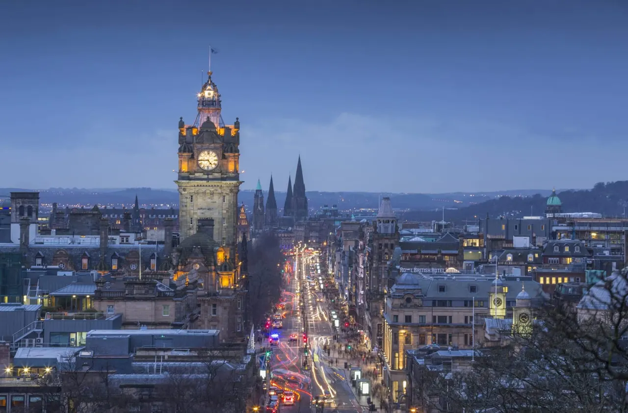 Illuminated clock tower dominates Edinburgh's historic cityscape at dusk, overlooking busy street with vehicle lights and classical architecture.