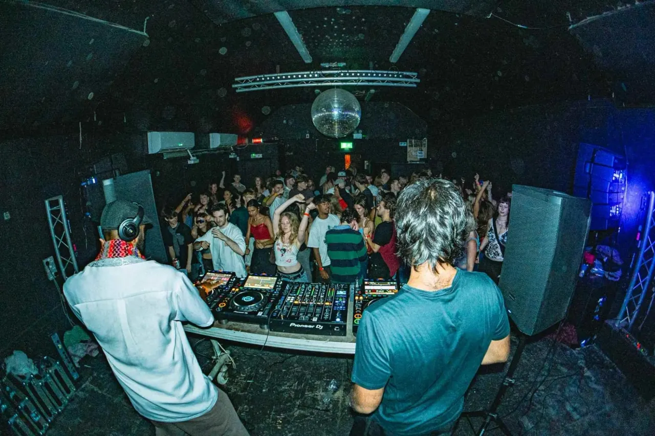 DJs behind decks entertaining a crowded dance floor in a dark nightclub with a disco ball overhead.
