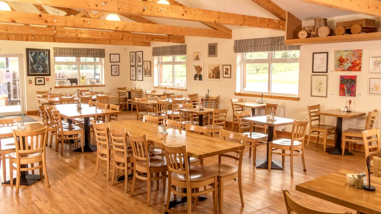 Spacious dining room with wooden tables and chairs, exposed beam ceiling, large windows overlooking fields, and countryside artwork on walls.