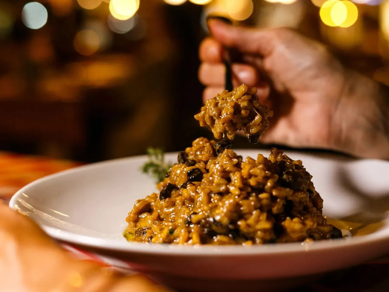 Person lifting a forkful of golden risotto from a plate in a warmly lit dining setting.