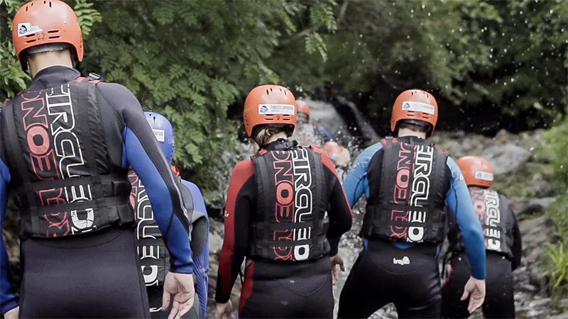 Group of people wearing orange helmets and wetsuits with life jackets standing by a river surrounded by trees.