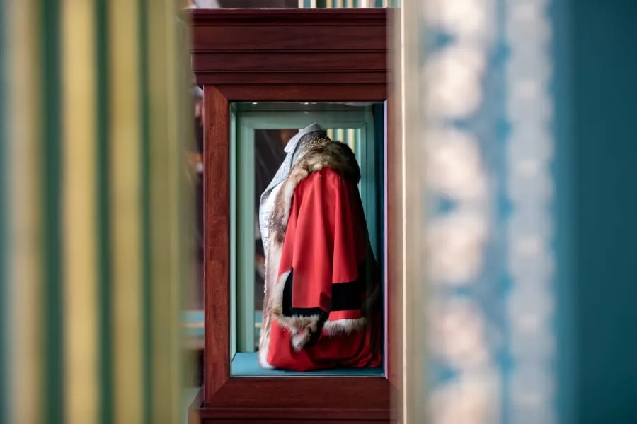 A dressed figure wearing a red coat trimmed with white fur and a white hat displayed in a glass case within a wooden frame.