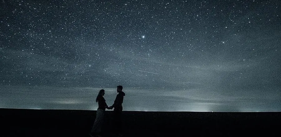 Two people holding hands stand silhouetted against a starry night sky with the Milky Way visible.