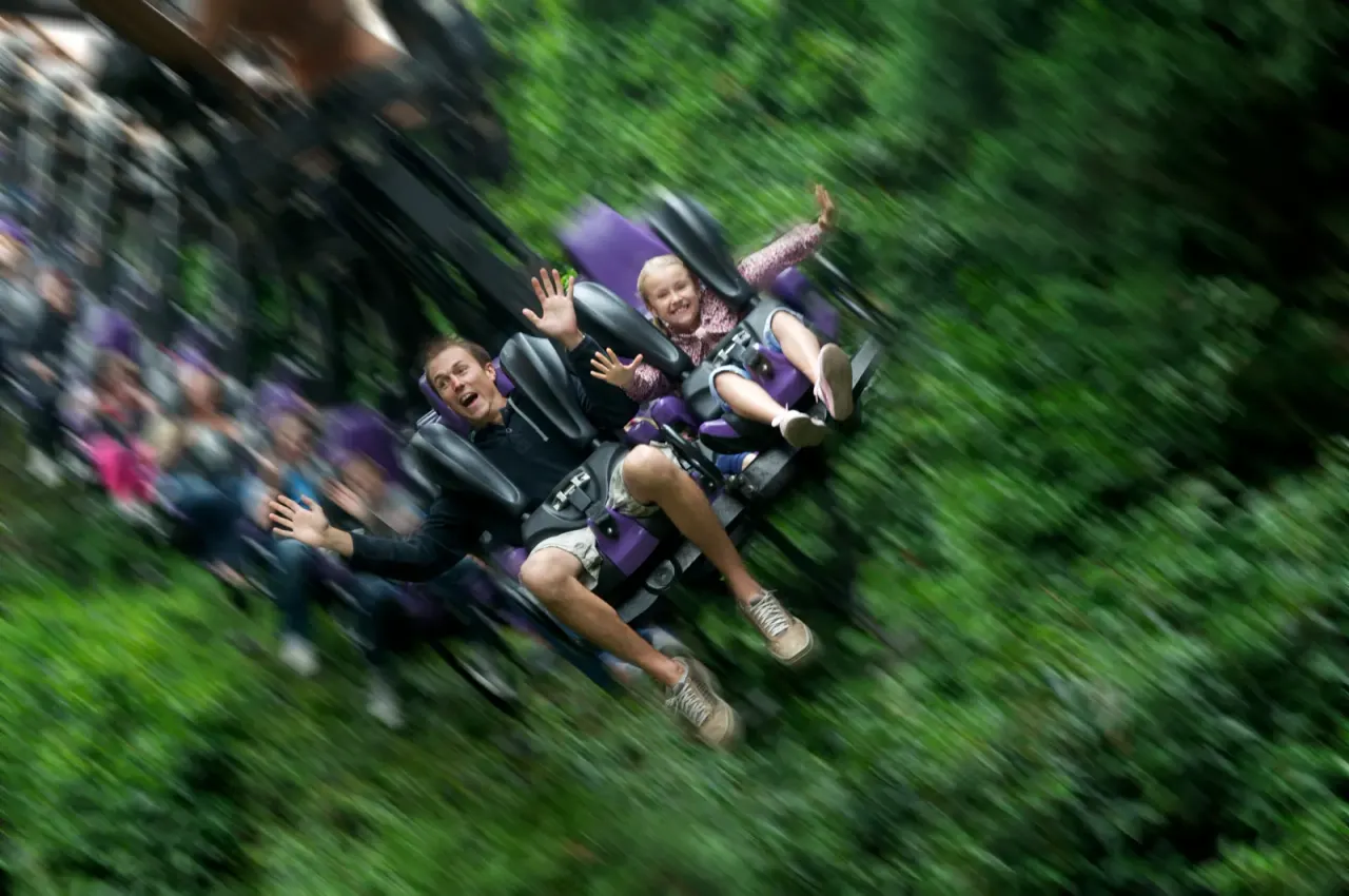 Three children on a theme park ride moving rapidly through green foliage, with excited expressions and raised arms.
