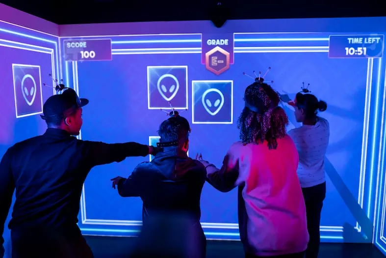 Four people play an interactive alien-themed video game on a large illuminated screen in a neon-lit gaming booth.