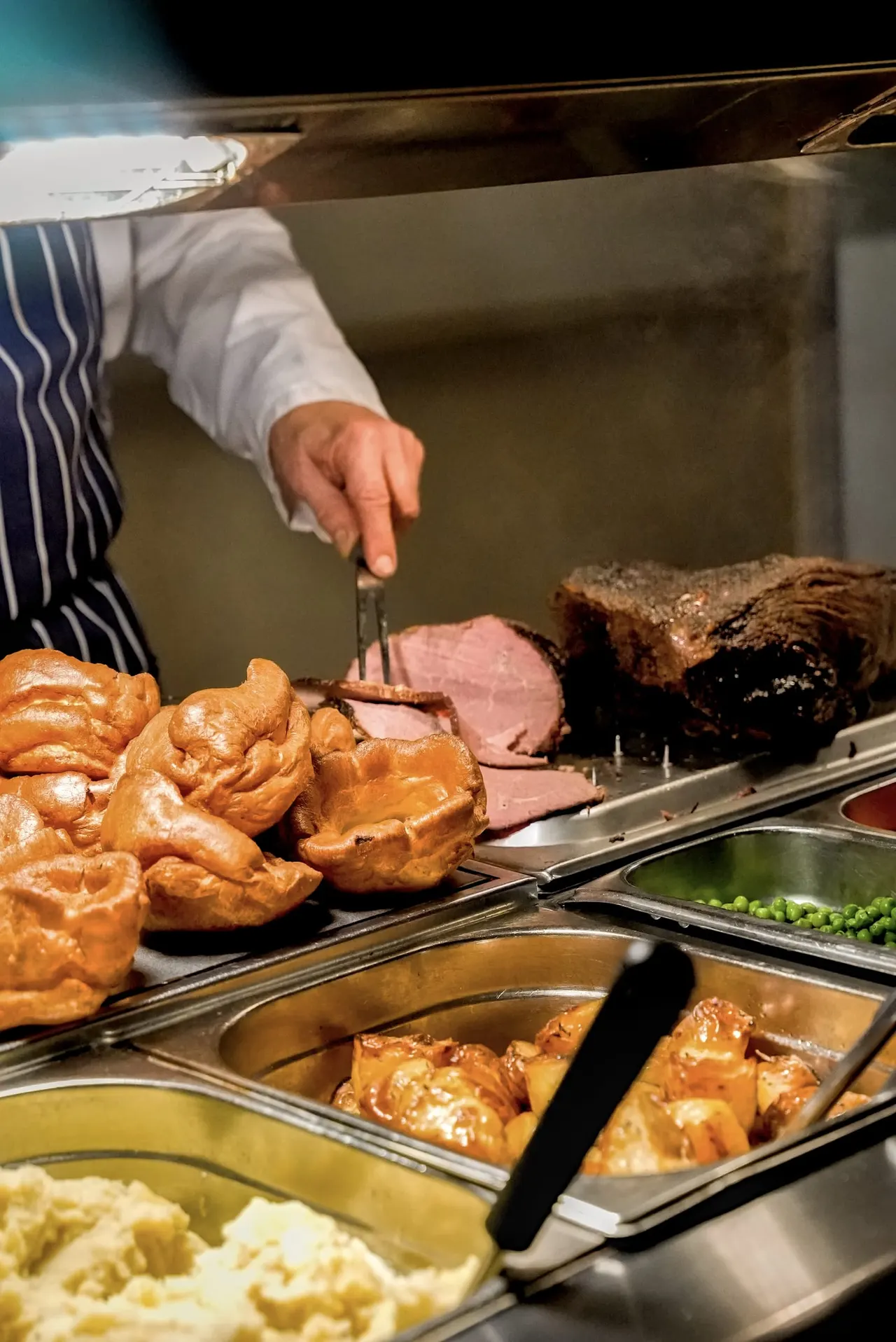 Server carving roasted meat at a carvery buffet with Yorkshire puddings, vegetables and other sides.