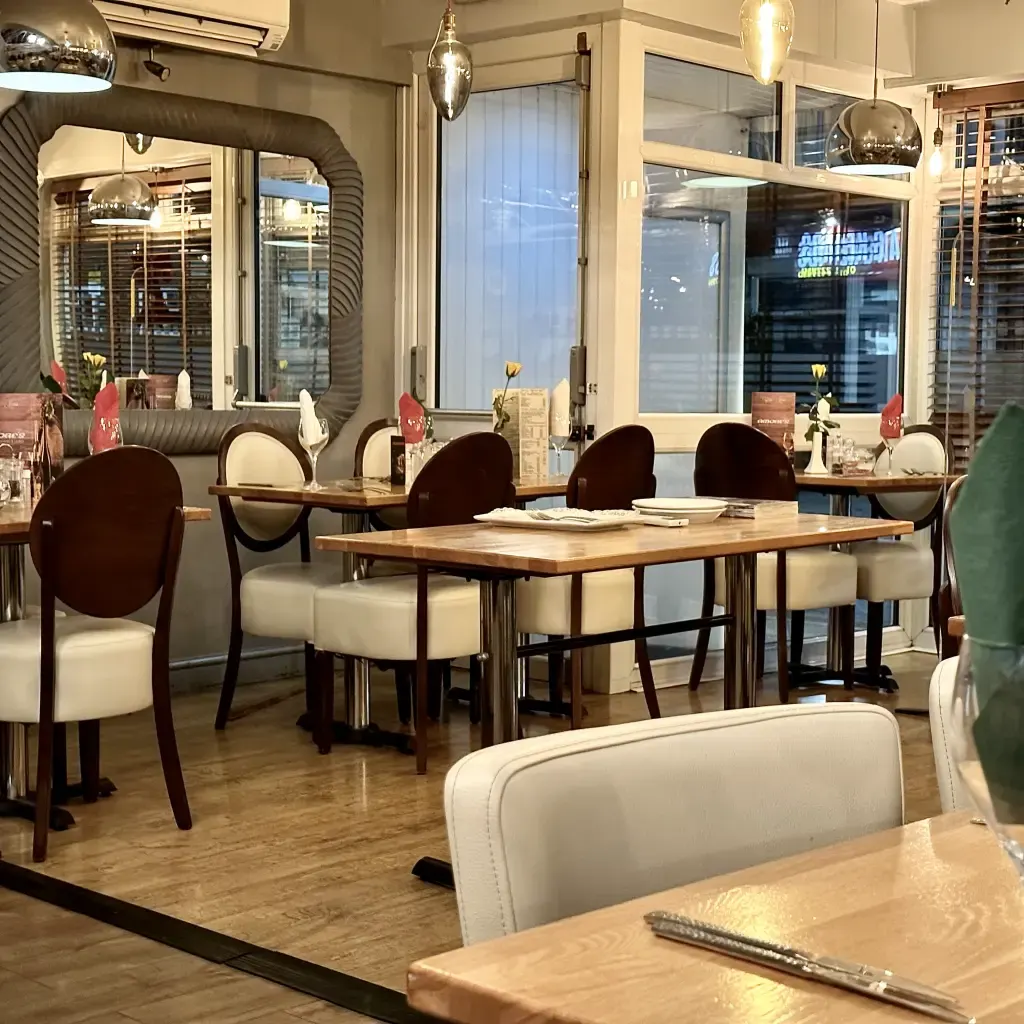 Modern restaurant dining room with wooden tables, cream and burgundy chairs, and large windows overlooking the city at night.