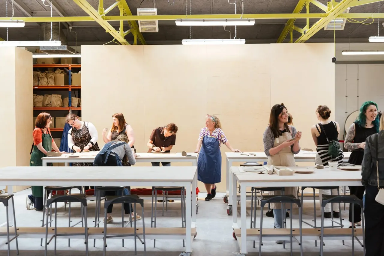 A group of participants engaged in a pottery or ceramics workshop at long work tables in an industrial studio space.