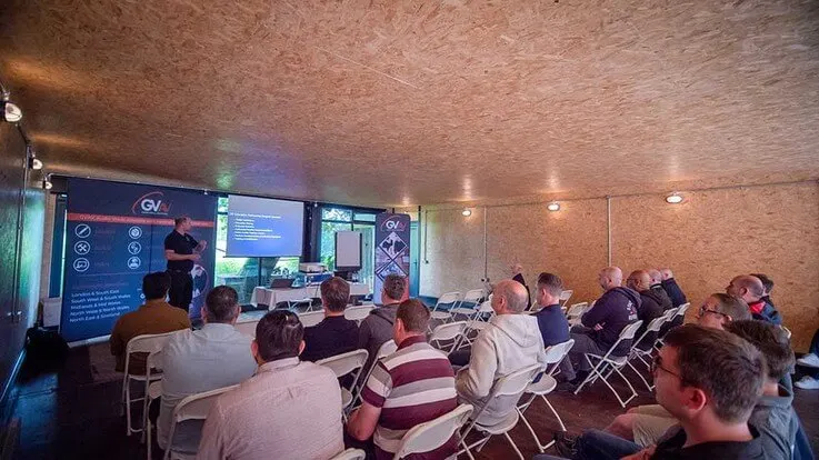 A presenter addresses an audience seated in chairs inside a brick-walled indoor venue, with digital screens displaying information behind them.