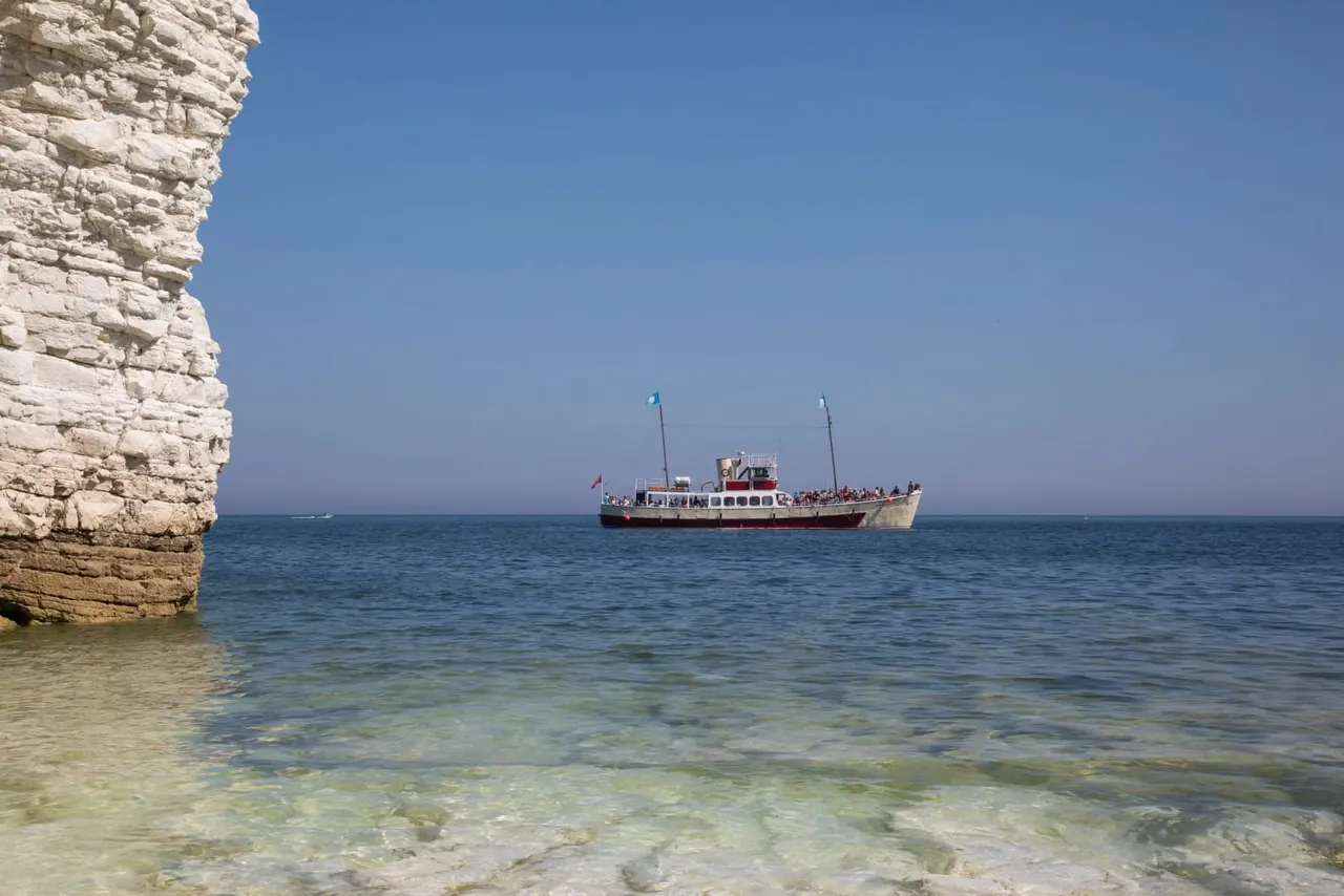 Passenger boat moored offshore next to white chalk cliffs in clear shallow water under blue sky.