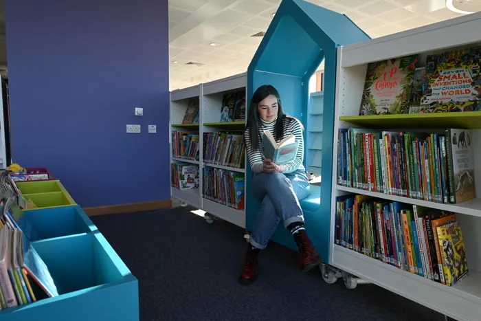 Young person sitting inside a turquoise book-shaped reading nook in a library surrounded by shelves of books.