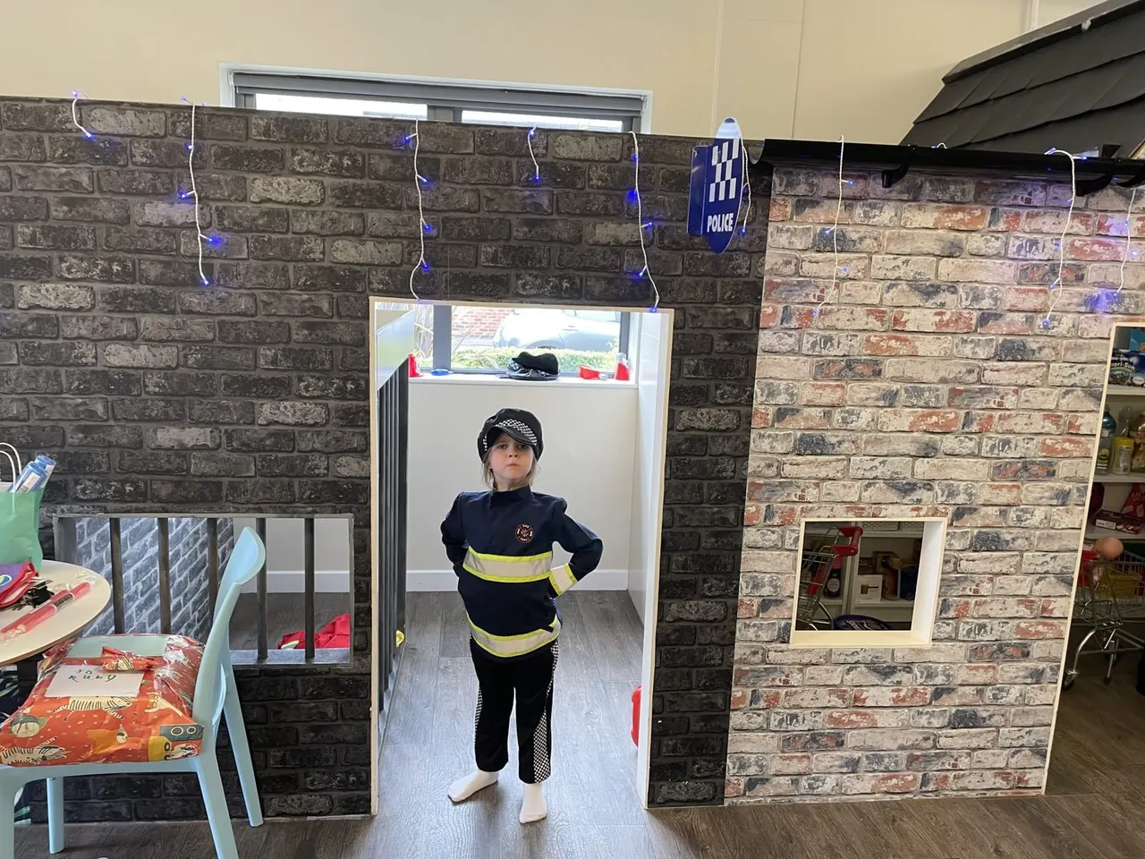 Child in police officer fancy dress standing in a decorated play area with brick-effect walls and blue fairy lights.