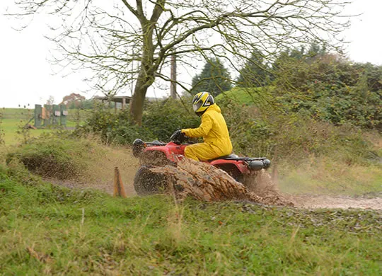 Person in yellow jacket riding a quad bike through muddy terrain at an outdoor activity venue.