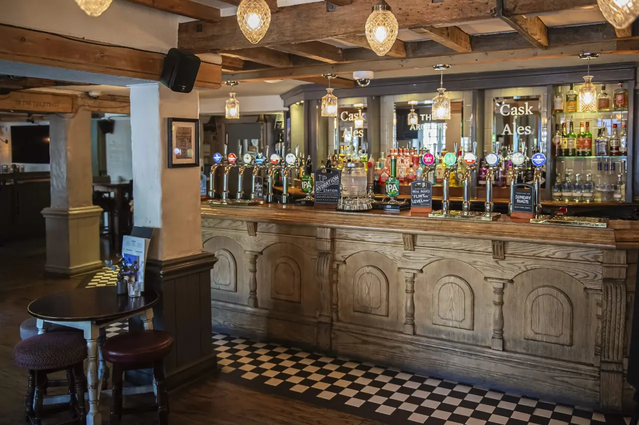 Interior of a traditional pub with a long wooden bar fitted with multiple beer pumps, shelved spirits behind, and a checkered floor.