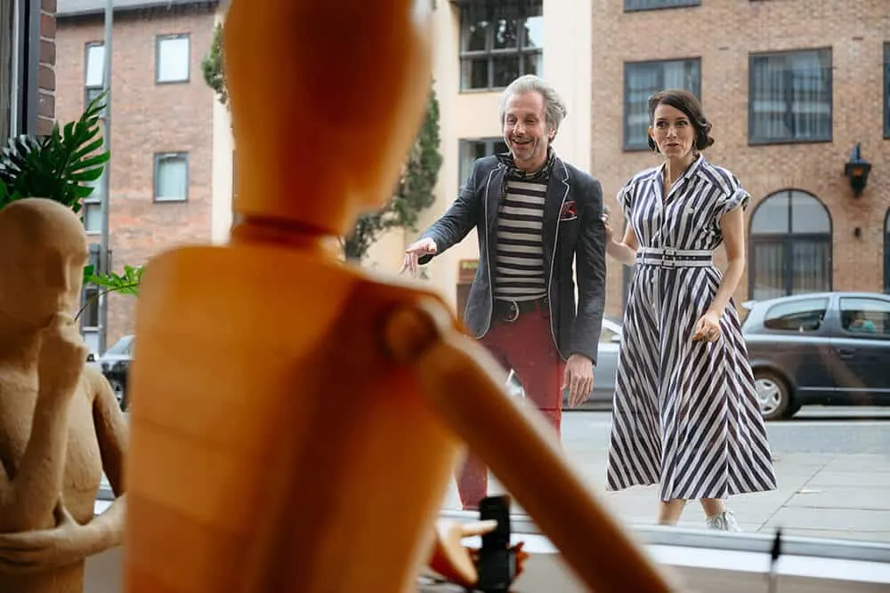 A man and woman smile while walking past mannequins displayed in a shop window on a city street.