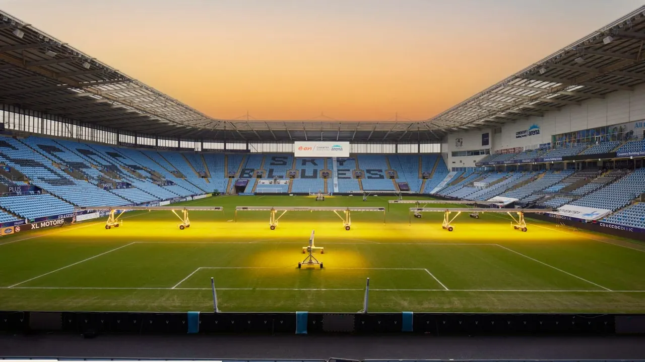 Football pitch with pitch lighting equipment during golden hour at a large stadium with blue seating.