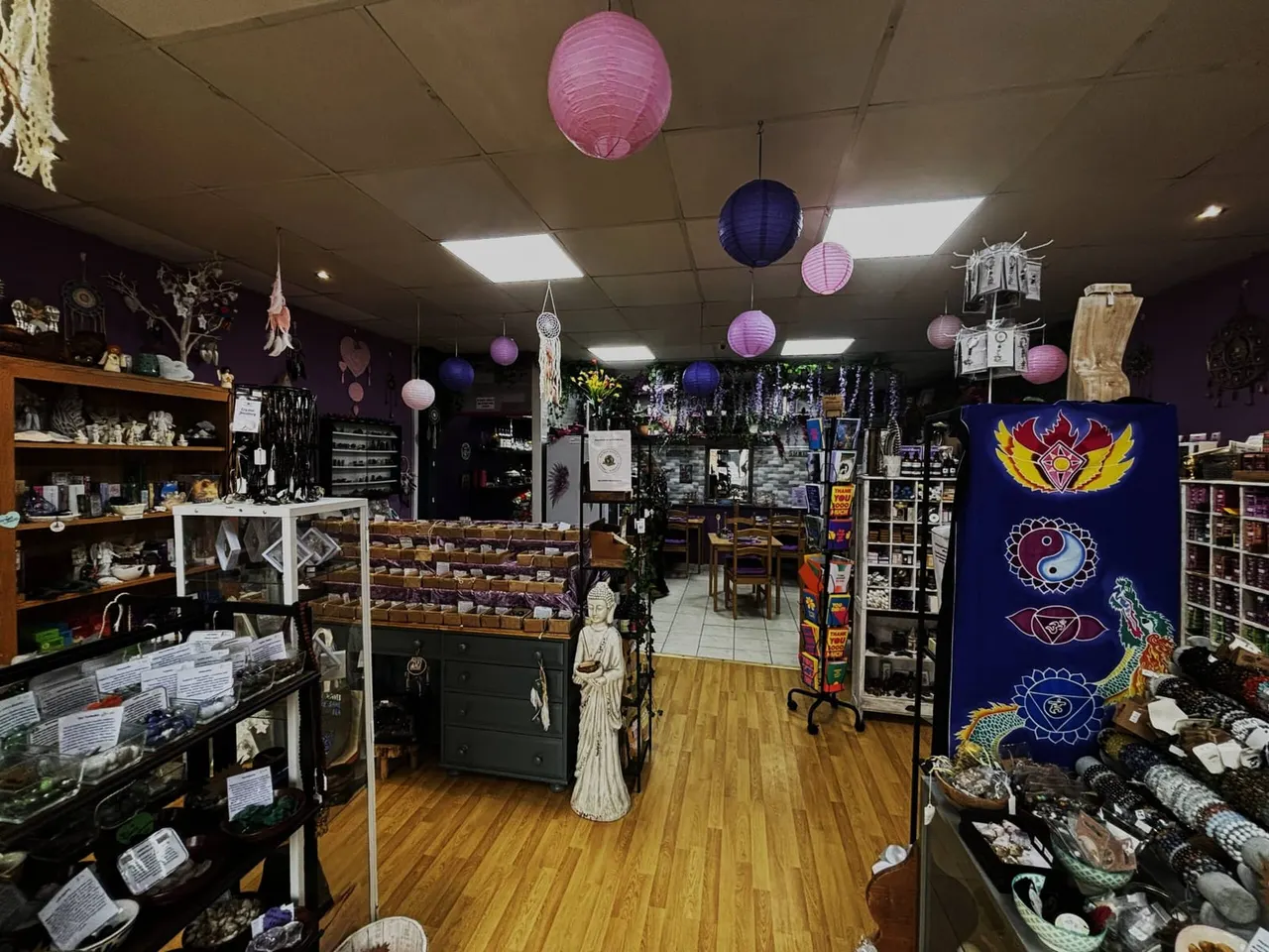 Interior of a New Age shop with purple walls, wooden floors, colourful paper lanterns, display cabinets of crystals and spiritual items, and decorative artwork.