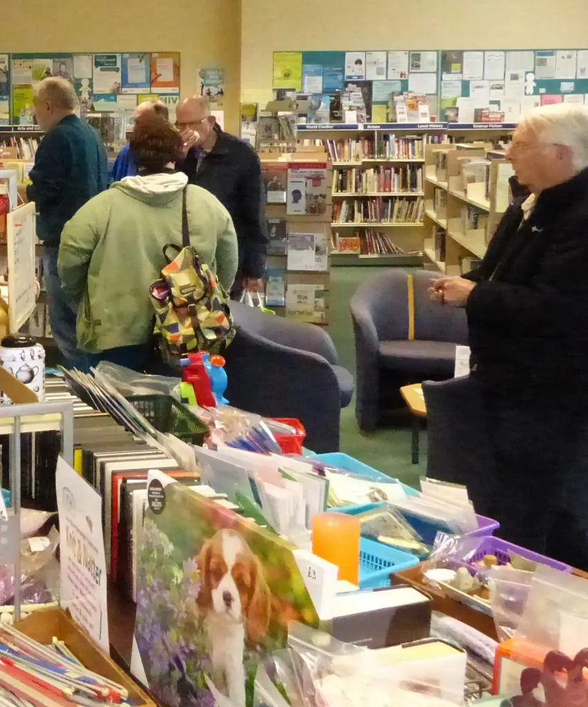 Adults browsing books and materials in a library with shelves and a display board in the background.