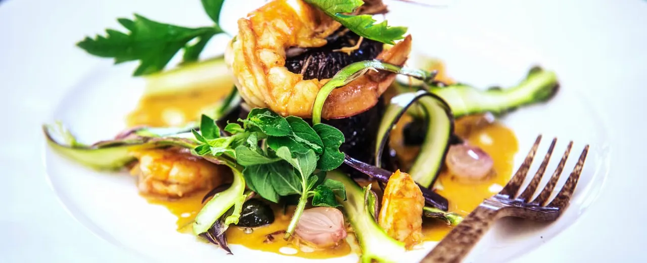 Plated fine dining dish with seared scallops, courgette ribbons, dark leaves and yellow sauce garnished with fresh herbs.