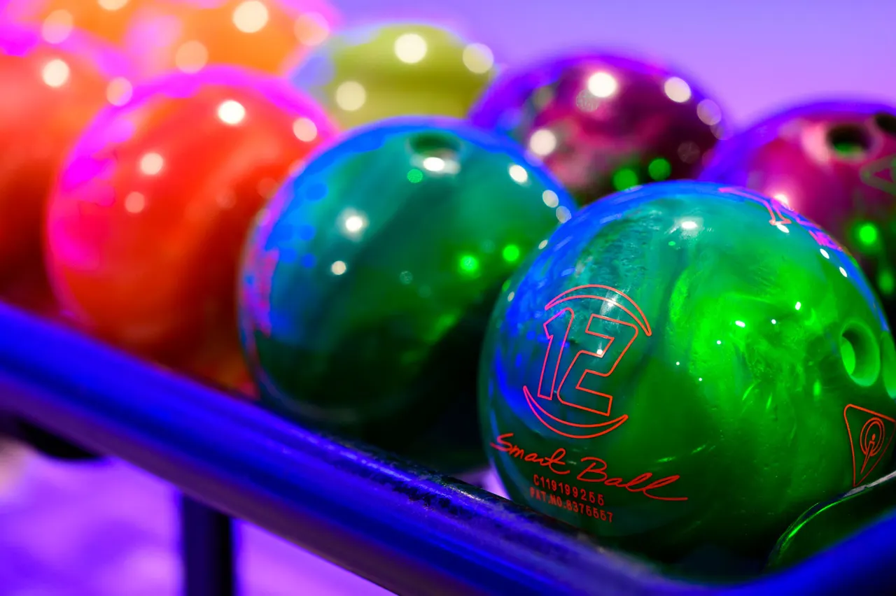 Colourful bowling balls lined up on a rack, illuminated by vibrant neon lighting in shades of pink, purple, green and blue.