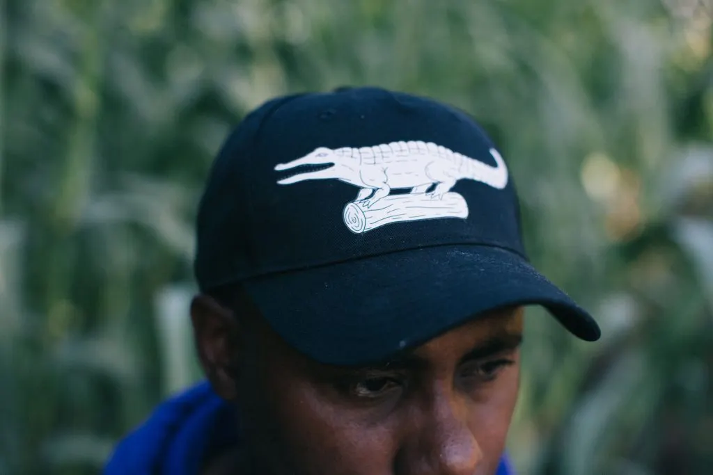 Person wearing a navy cap with a reptile logo against a blurred green foliage background.