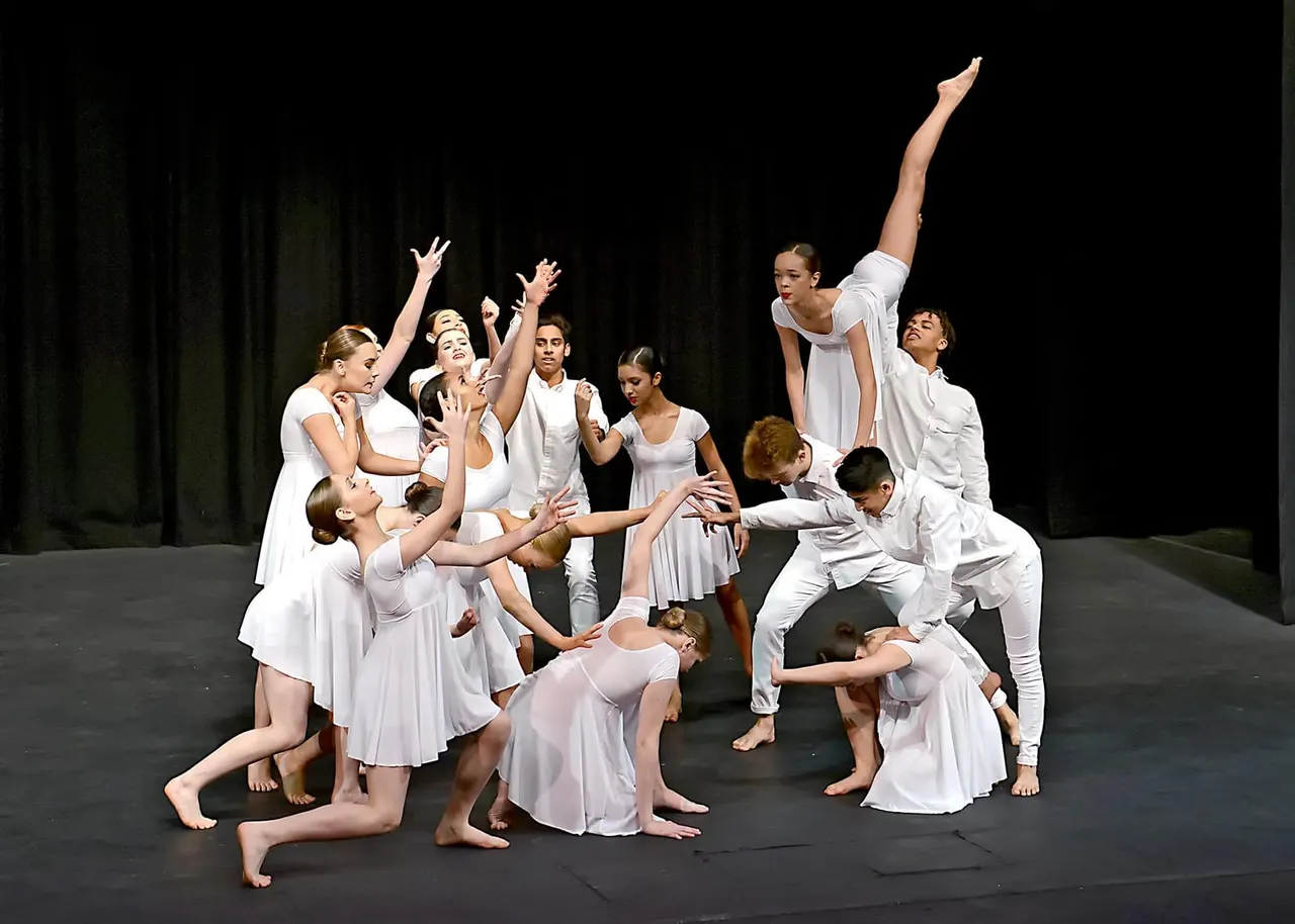 A group of young ballet dancers in white costumes perform together on a dark stage, with one dancer in an elevated arabesque pose.