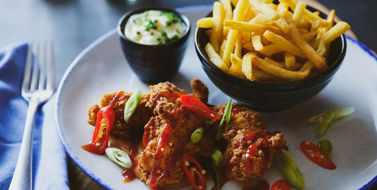 Fried chicken pieces with red peppers and spring onions served with chips and a creamy dip on a white plate.