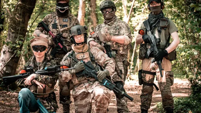 Group of people in tactical gear and protective masks holding paintball guns in a wooded area.
