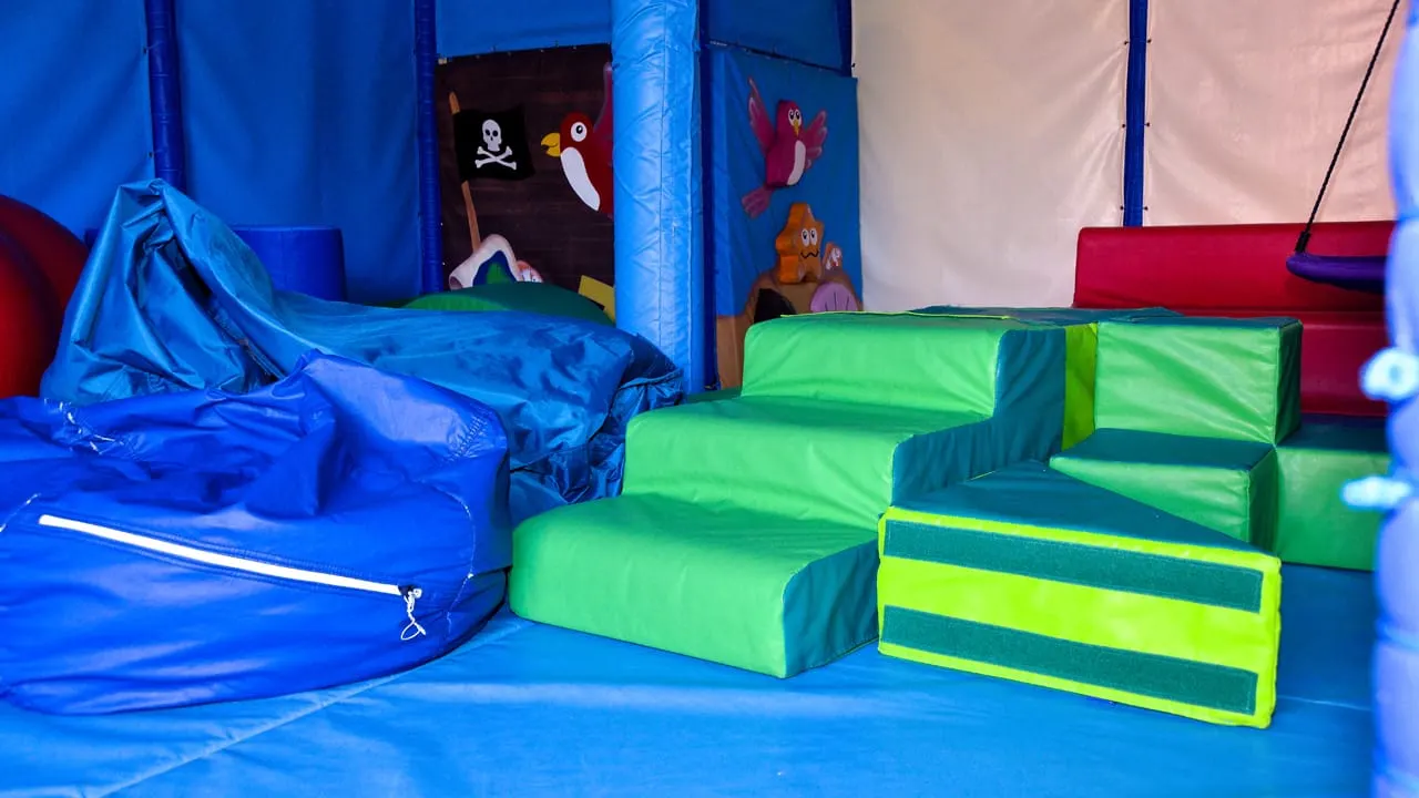 Interior of a soft play area with blue inflatable structures, green foam climbing blocks, and themed wall panels with cartoon characters.