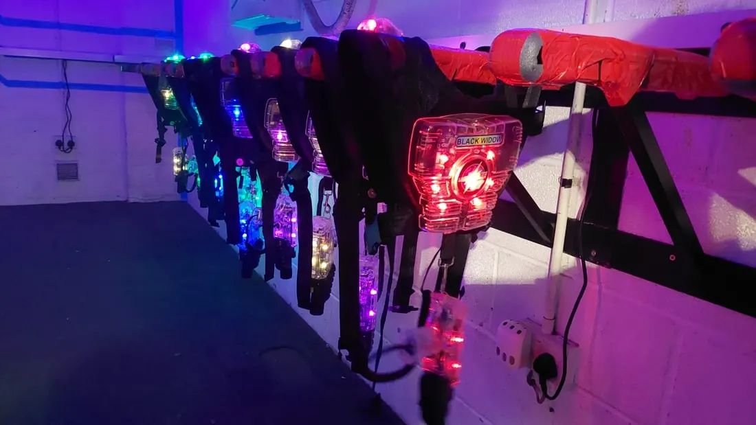 Row of illuminated laser tag guns and equipment under purple and blue lighting in an indoor arena.