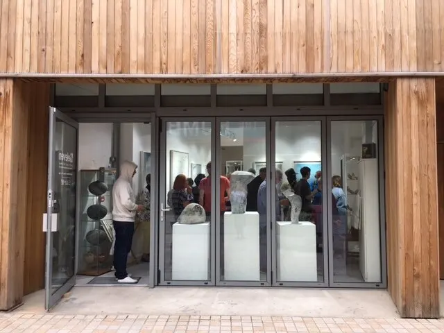Visitors standing inside a modern gallery space with large glass doors and wooden exterior cladding.