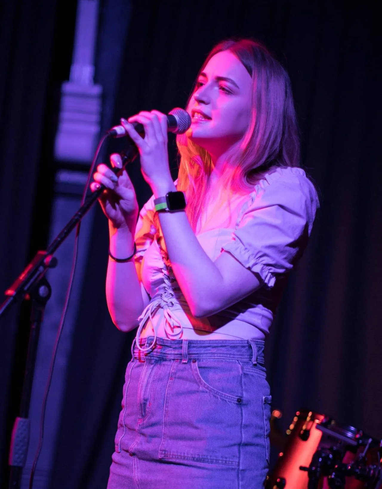 A young woman singing into a microphone on stage, bathed in pink and purple stage lighting.