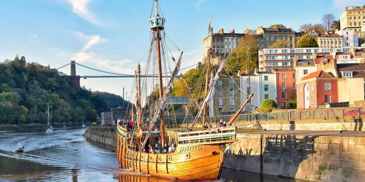 Historic replica sailing ship moored at Bristol Harbourside with colourful Georgian buildings and suspension bridge in background.