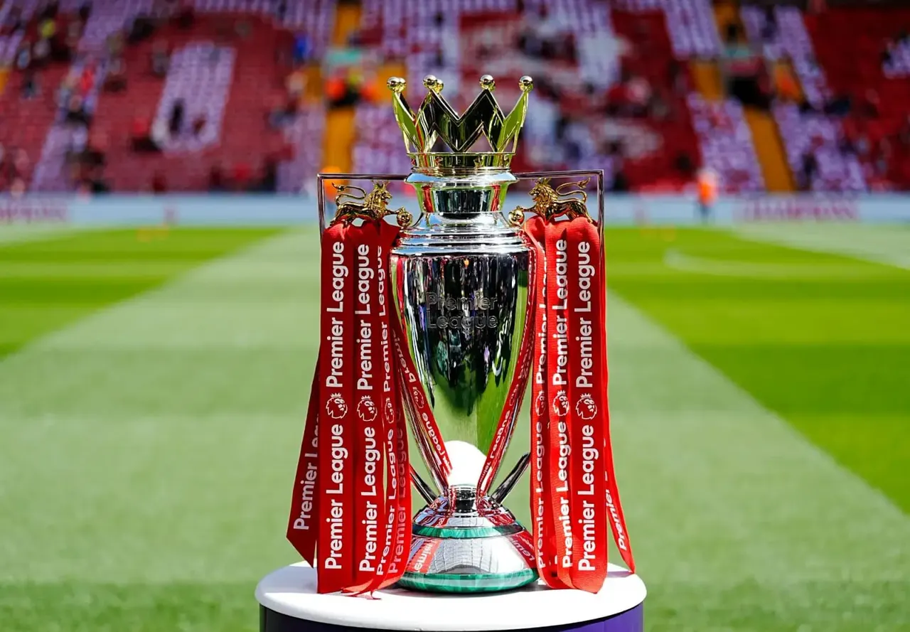 Premier League trophy with crown-topped silver vessel and red ribbons displayed on a football pitch inside a stadium.