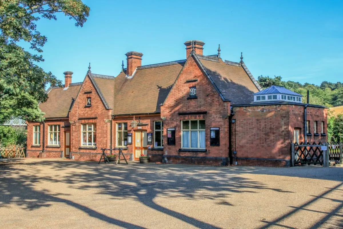 Victorian brick railway station building with pitched tiled roofs and chimneys under clear blue sky.