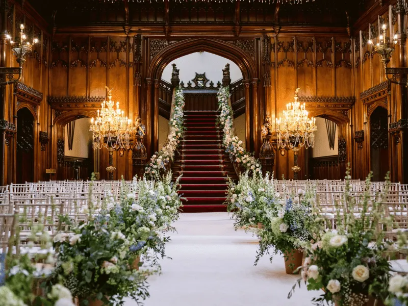 Ornate wooden hall with chandeliers, white chairs lining an aisle decorated with white and blue flowers leading to a grand staircase.