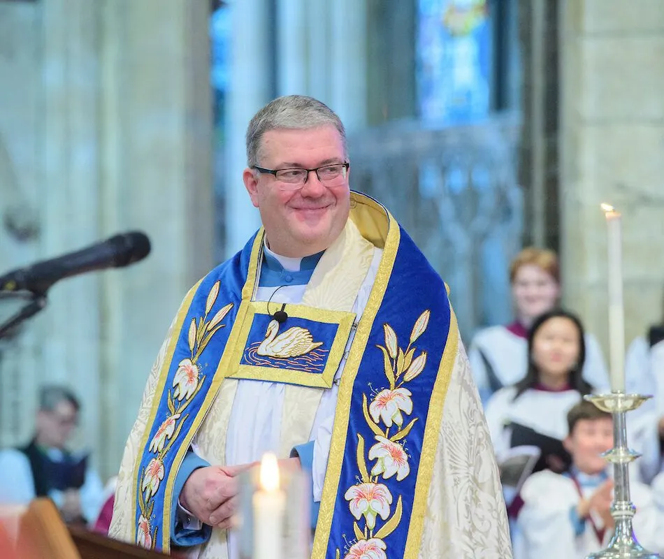 Clergy member in ornate blue and gold vestments holding a candle during a church service.
