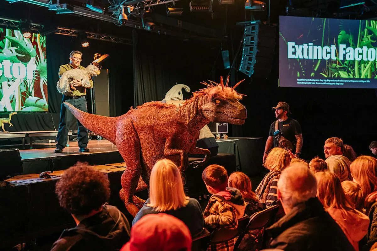 Presenter on stage with a large red dinosaur model demonstrating to an audience of children seated in front, with screens showing "Extinct Factor" content.