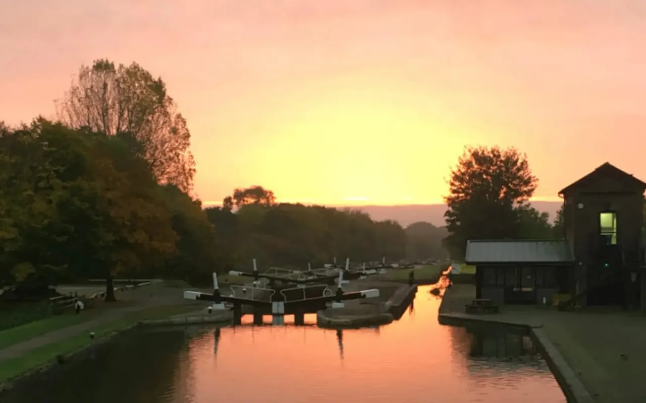 Canal lock system at sunrise with moored narrowboats, a lock keeper's cottage, and surrounding woodland reflected in still water.