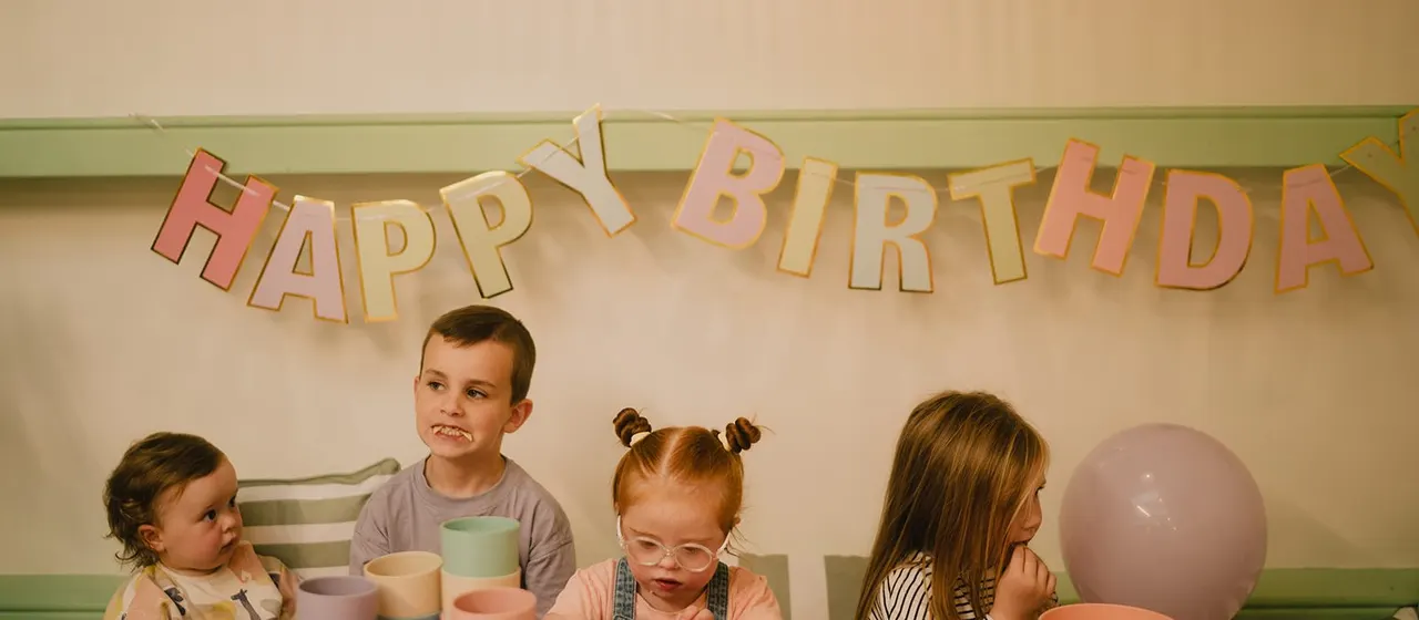 Four young children seated at a table under a "Happy Birthday" banner during a party celebration.