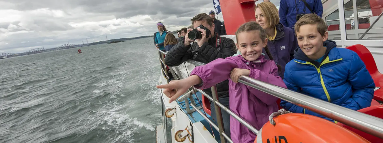 Children and adults on a boat tour leaning against the rail, with one person photographing the seascape and industrial coastline.