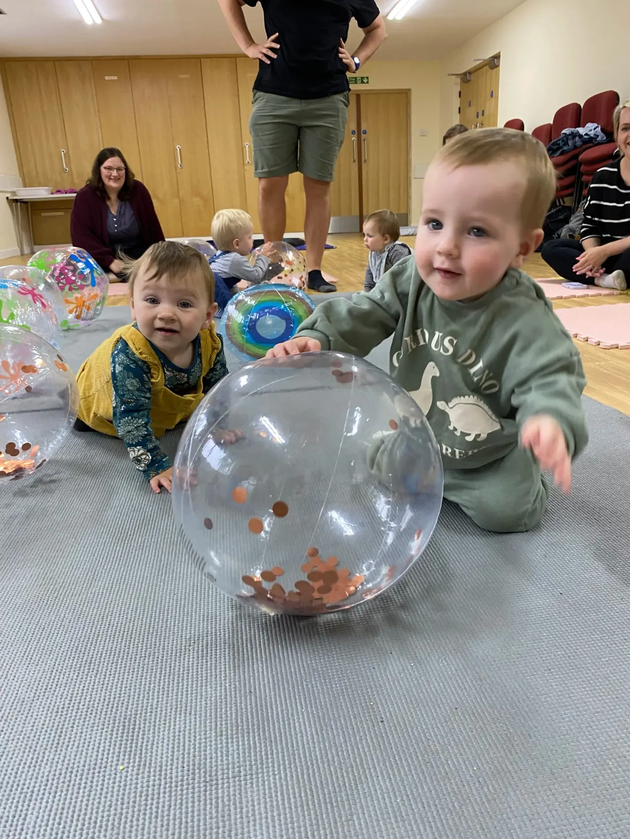 Toddlers play with transparent balloons filled with coloured confetti during an indoor activity session with caregivers.