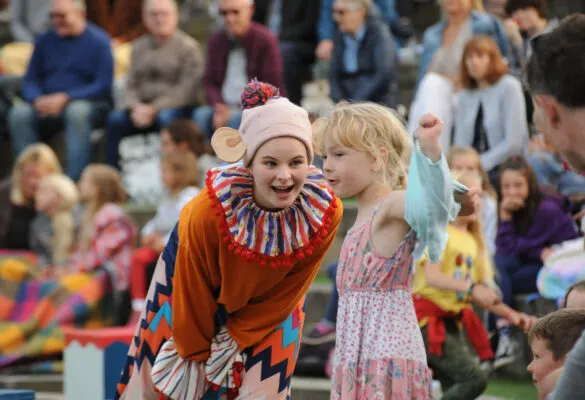 Child dressed as a colourful clown performs alongside another child whilst audience members watch from tiered seating.